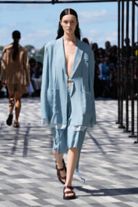 Model walks the runway in a light-blue oversized blazer and matching shorts, open at the chest, on a patterned floor.
