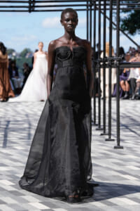 Model walks a black strapless gown down an outdoor runway, dramatic silhouette and sheer train trailing behind her.