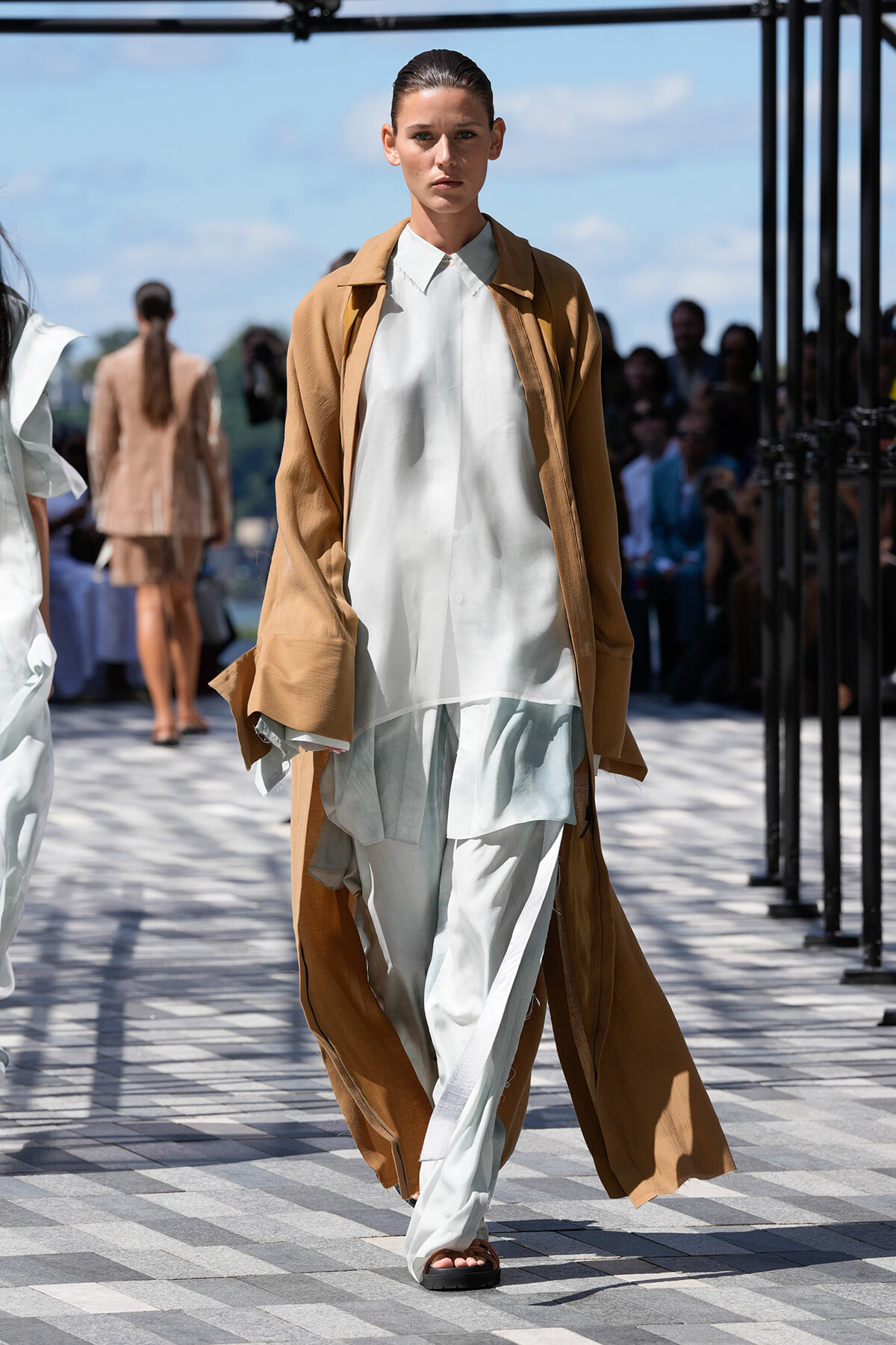 Model walking an outdoor runway in a camel long coat over a white, layered top and pale pants; audience blurred in background.