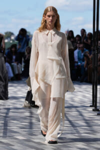Model walking a runway in a sheer, layered cream-colored outfit with sandals, outdoors with spectators in the background