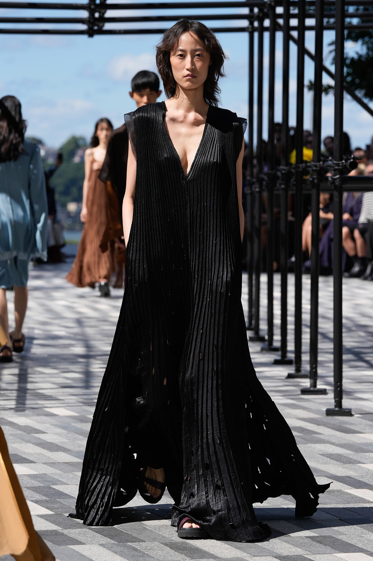 Model wearing a long black pleated gown with a deep V-neck walking down a runway.