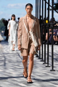 Model walks a fashion runway in a beige linen suit with white detailing, wearing flat tan sandals.