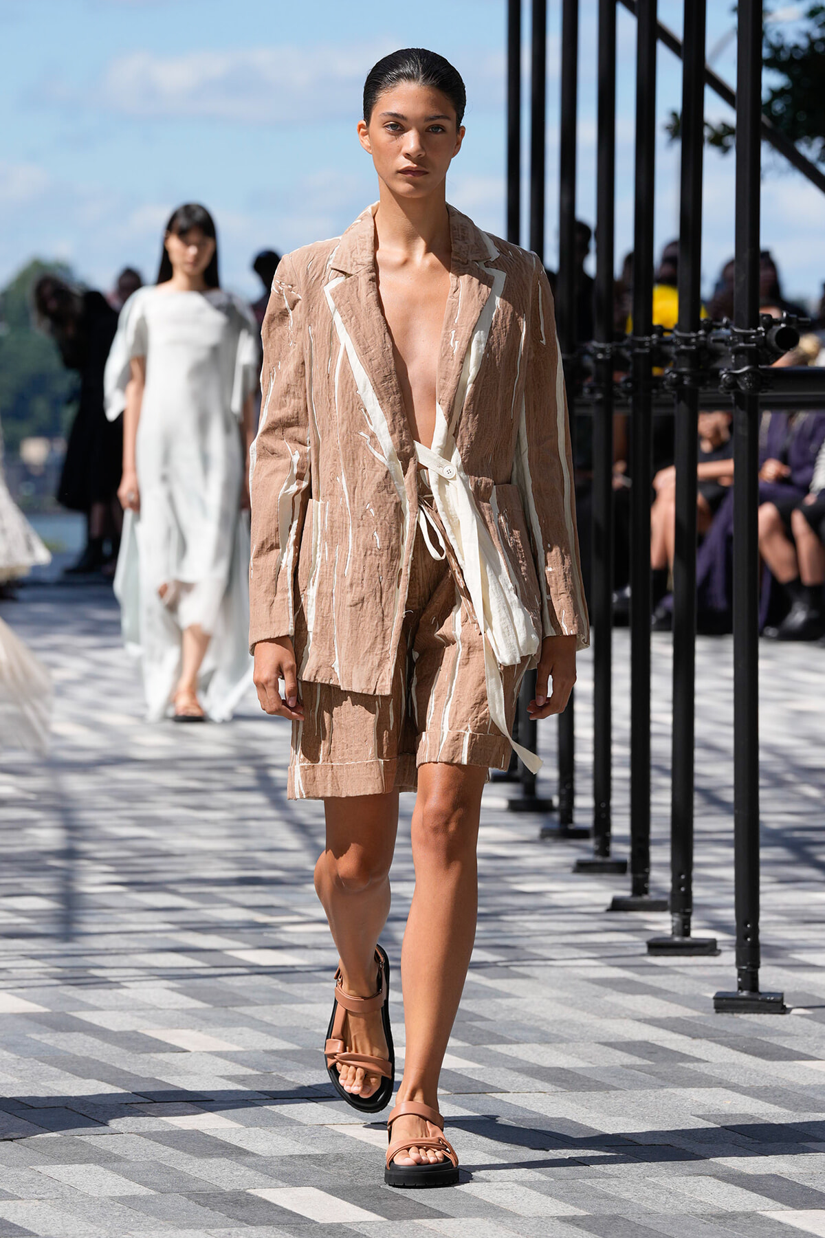 Model walks a fashion runway in a beige linen suit with white detailing, wearing flat tan sandals.