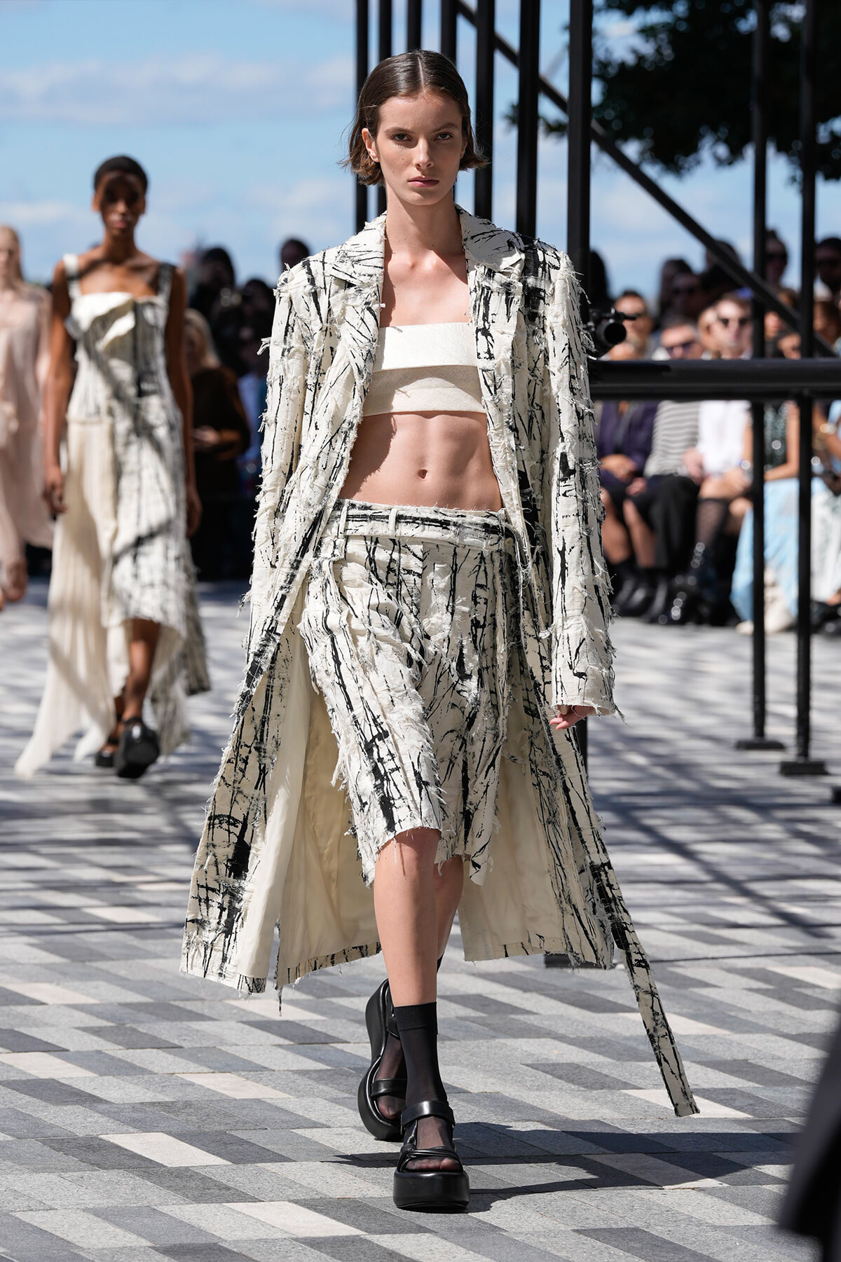 Model walks a fashion show wearing a cream textured long coat over a matching cropped bandeau and high-waisted shorts with a black abstract pattern, black platform sandals