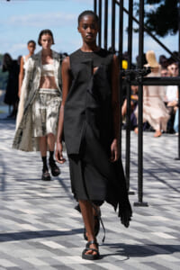 Outdoor fashion show runway with a dark-skinned model in a black dress walking toward the camera; others in stylish outfits behind her.
