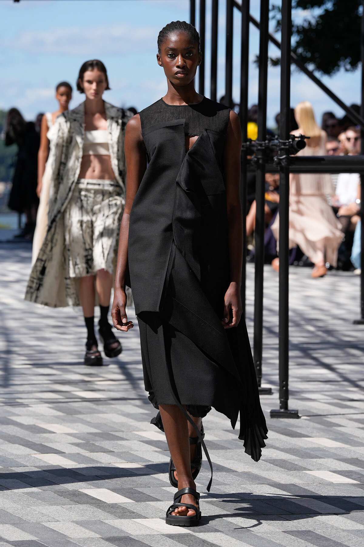 Outdoor fashion show runway with a dark-skinned model in a black dress walking toward the camera; others in stylish outfits behind her.
