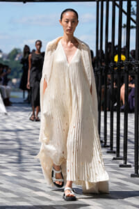 Model walking a runway in a cream pleated dress with a flowing cape and white sandals.