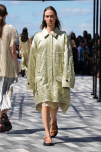 Model wearing a crinkled pale-green oversized coat with large pockets and tan sandals on an outdoor runway.