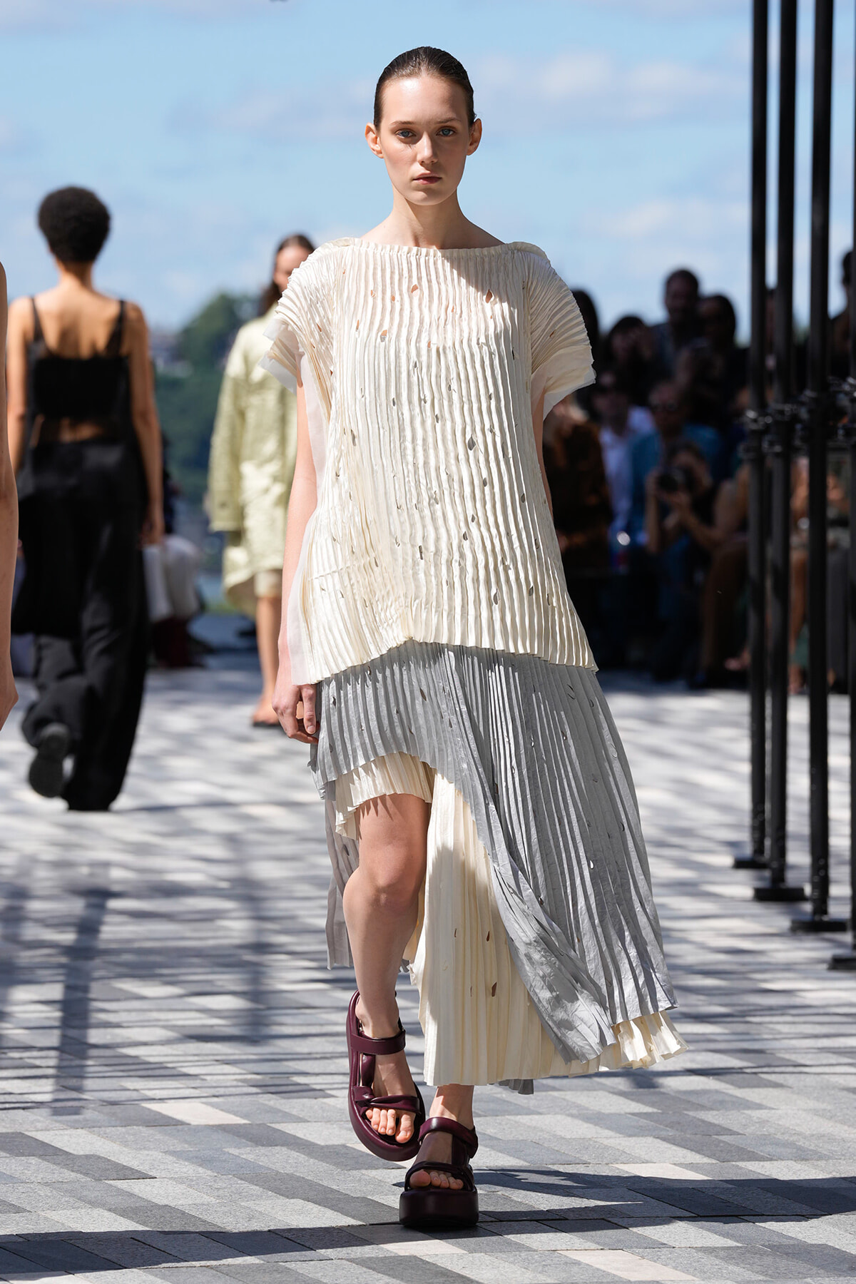 Female model on an outdoor runway wearing a cream pleated top with a layered gray skirt and burgundy platform sandals, walking toward the camera.