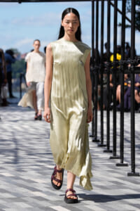 Model walking on an outdoor runway in a pale yellow pleated sleeveless dress with burgundy sandals; audience and railings in the background.