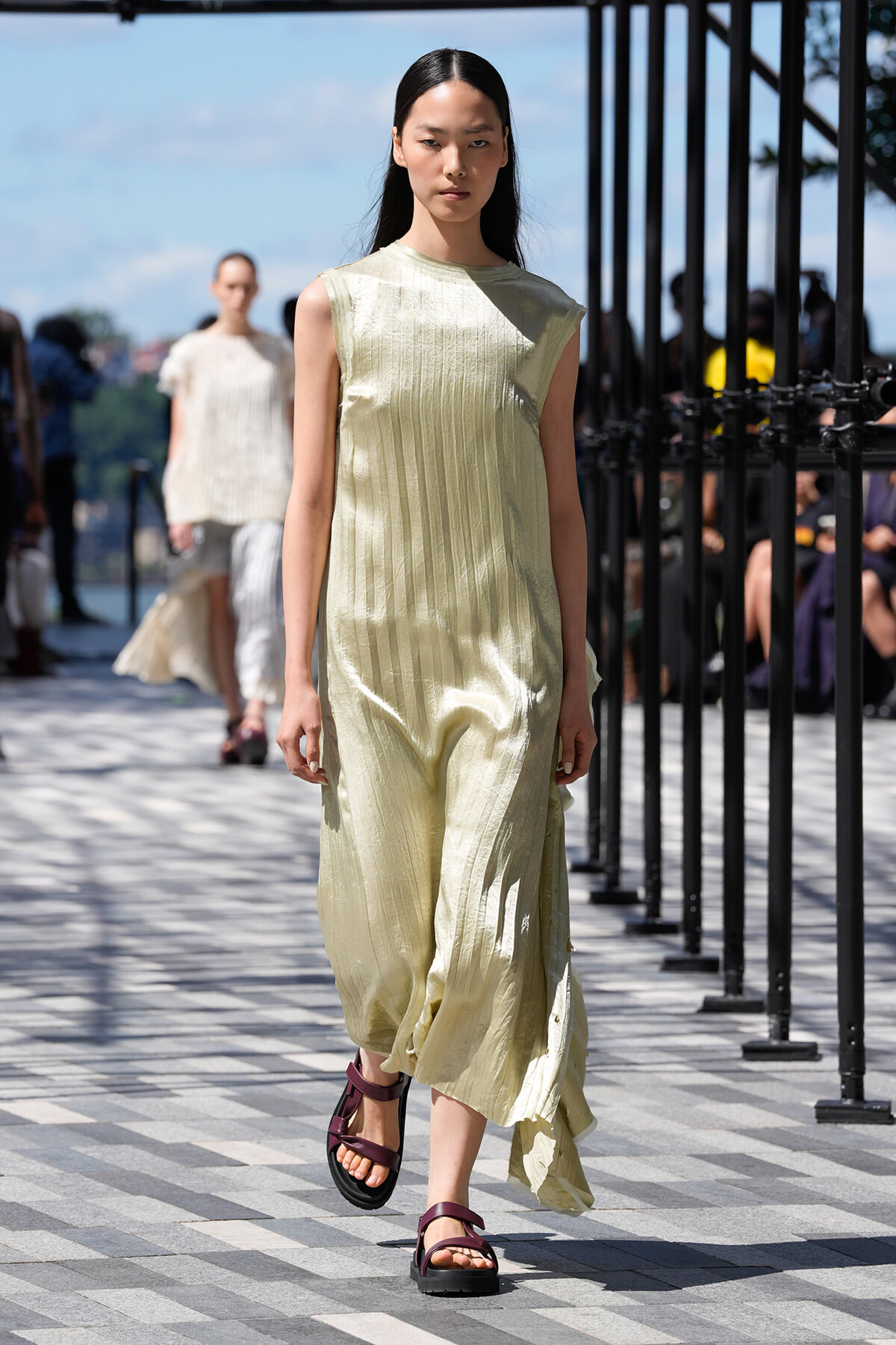 Model walking on an outdoor runway in a pale yellow pleated sleeveless dress with burgundy sandals; audience and railings in the background.