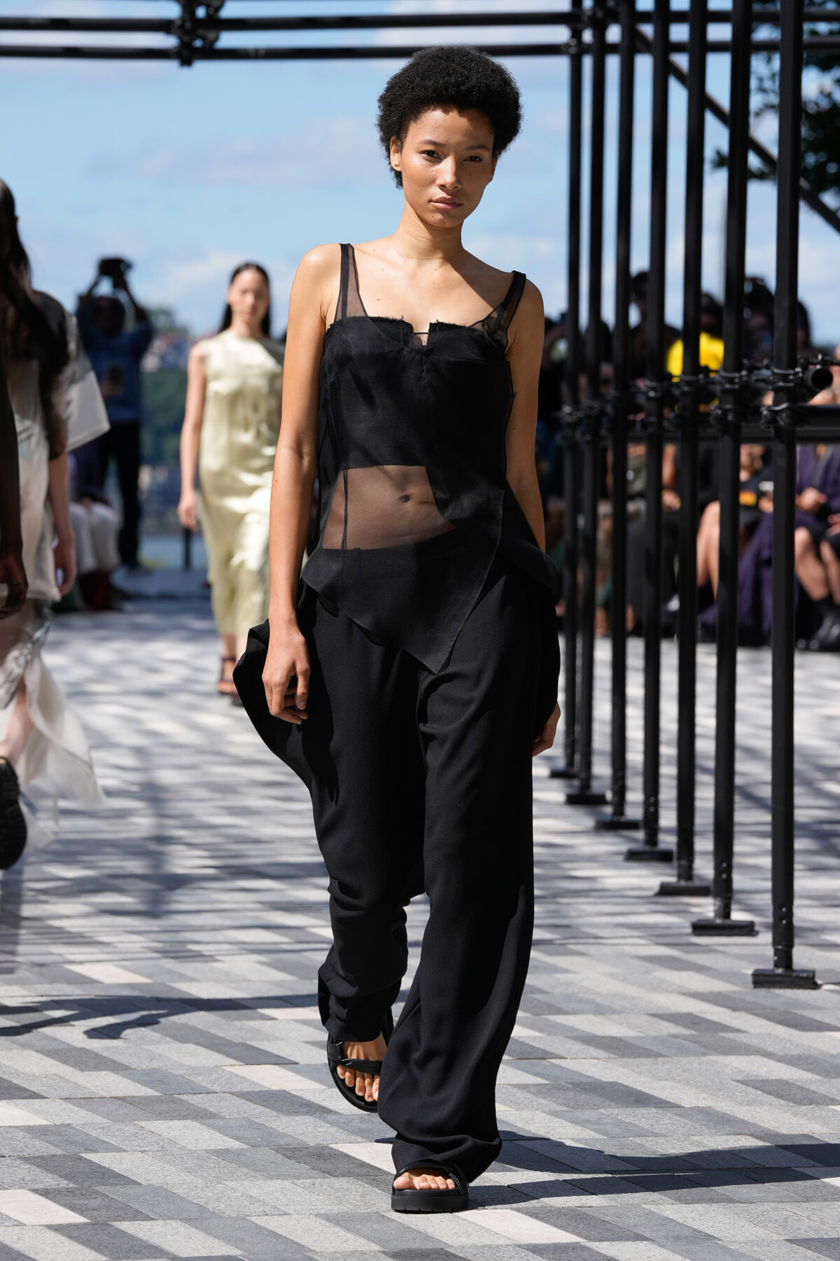 Model walks a black sheer-hemmed top and wide black pants on an outdoor runway, spectators in background.