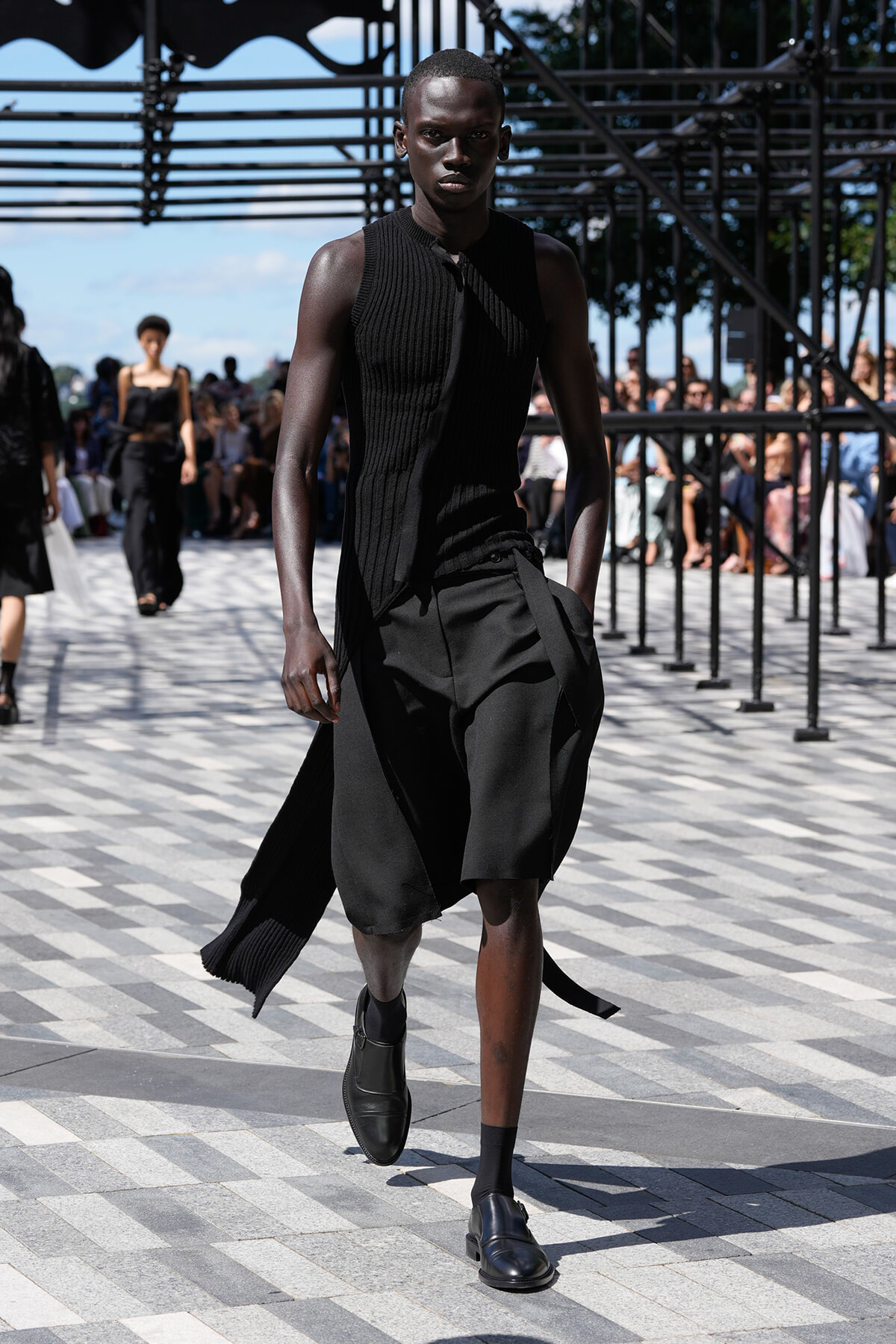 Male model walks a fashion show wearing an all-black sleeveless knit top and flowing knee-length shorts, with a crowd in the background.