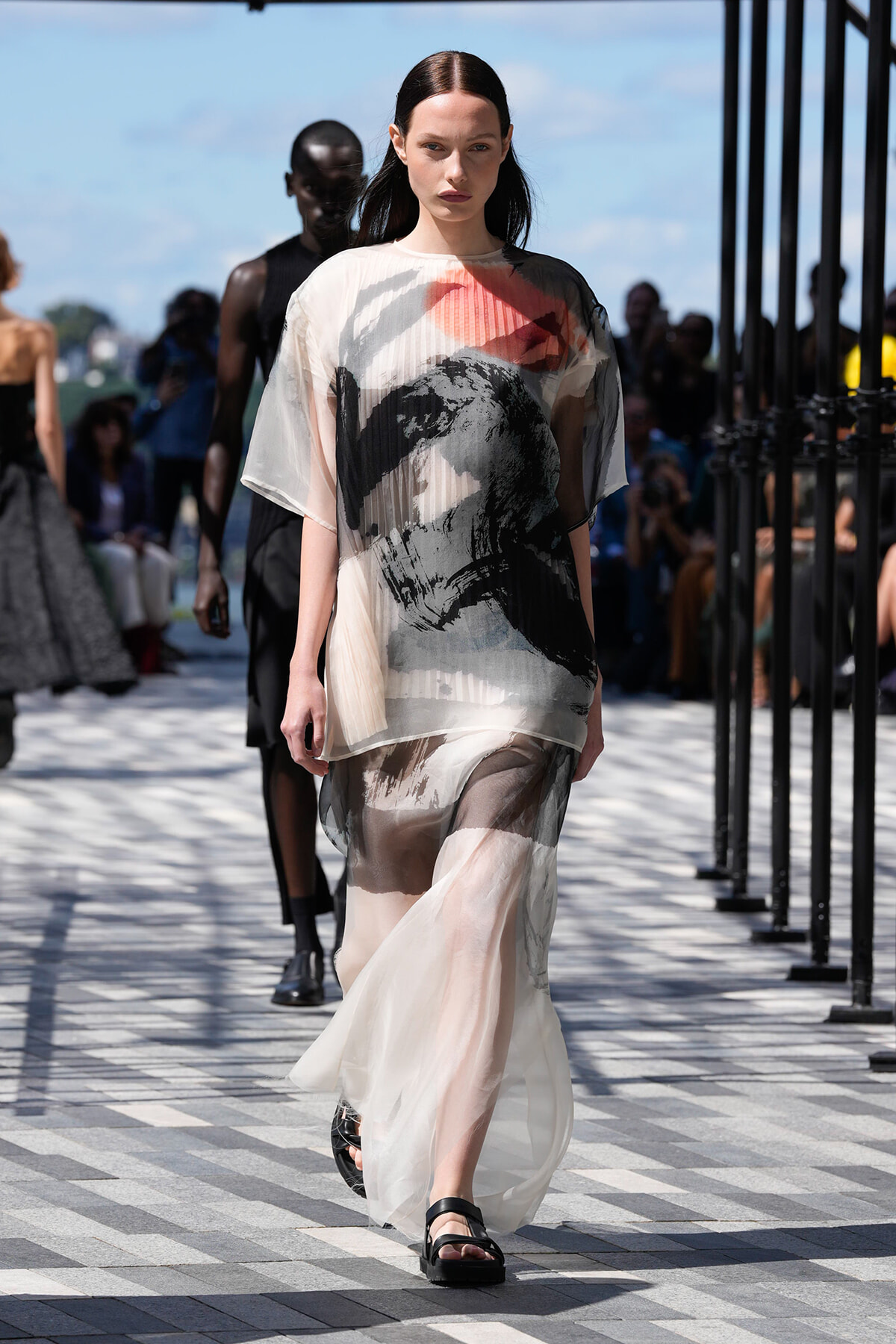 Model walks the runway in a sheer, abstract-printed dress with a flowing translucent skirt and black sandals