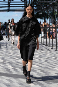 Model on an outdoor runway wearing a crinkled black top and black knee-length shorts, with chunky sandals; audience visible in the background.