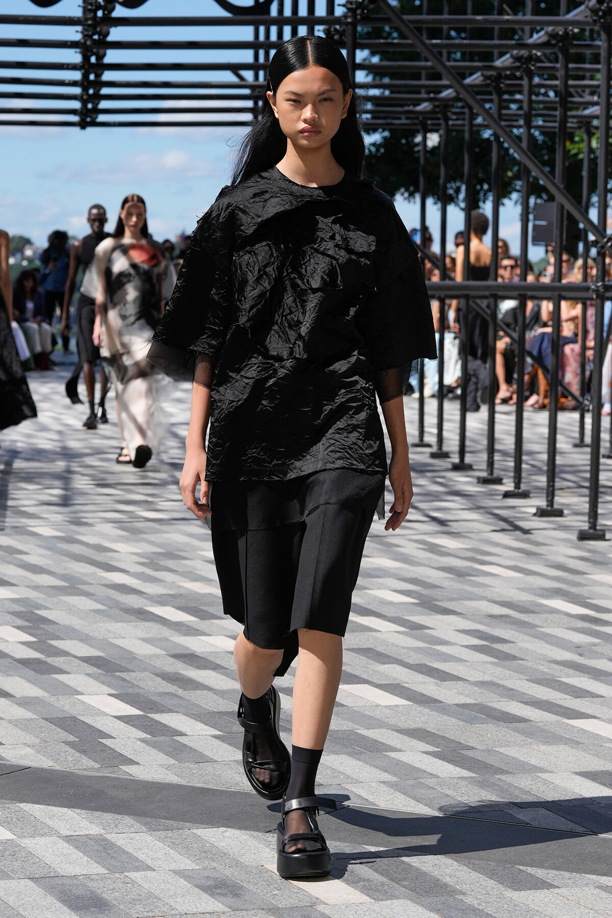 Model on an outdoor runway wearing a crinkled black top and black knee-length shorts, with chunky sandals; audience visible in the background.