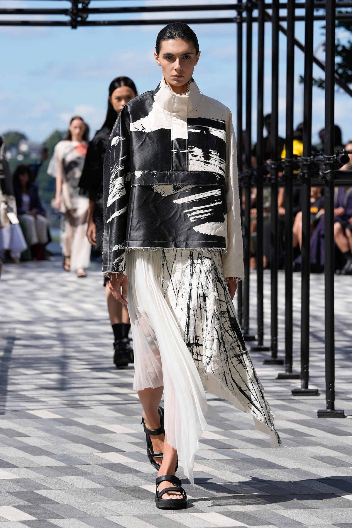 Model on a runway wearing a black-and-white abstract print top over a sheer white skirt, outdoors.