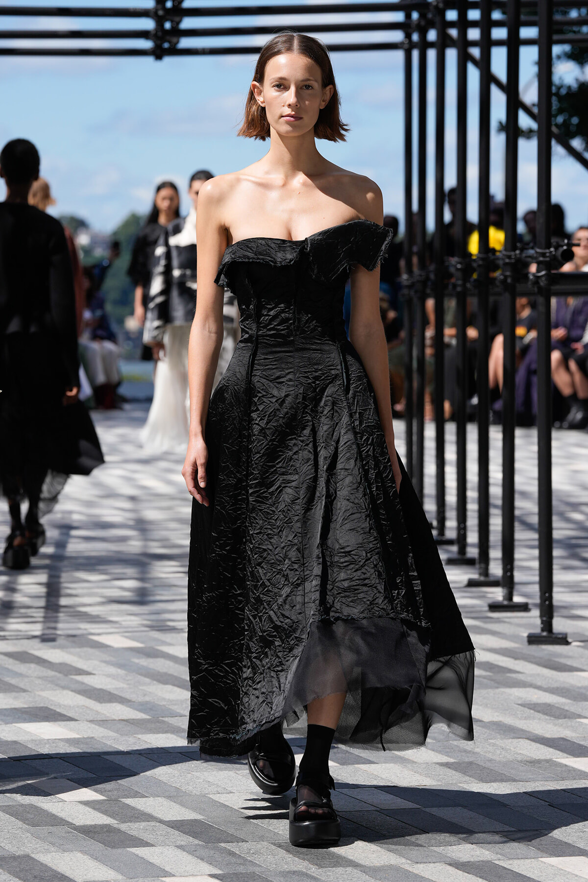 Model walking a runway in a black off-the-shoulder, crinkled maxi dress with a sheer hem under a metal framework on a sunny day.
