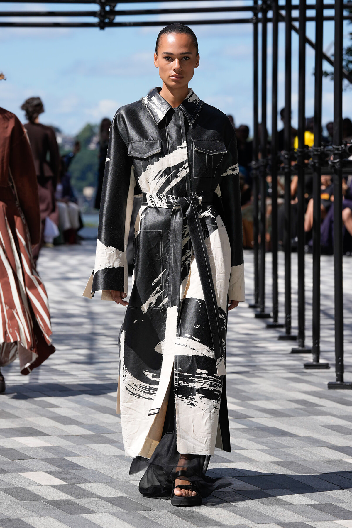 Model walks the runway in a long black-and-white abstract-print coat tied at the waist.