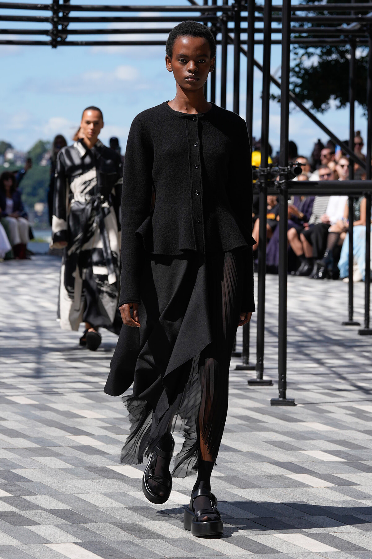 Model walking a fashion runway in an all-black ensemble with a structured, layered top and asymmetrical skirt with sheer panels outdoors.