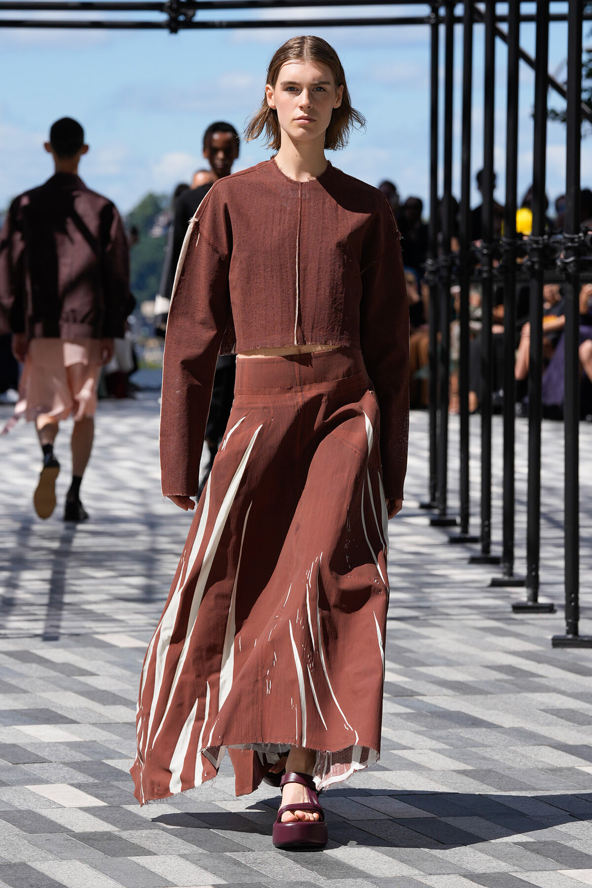 Model walking on an outdoor runway in a rust-colored cropped top and a long brown skirt with white abstract stripes.