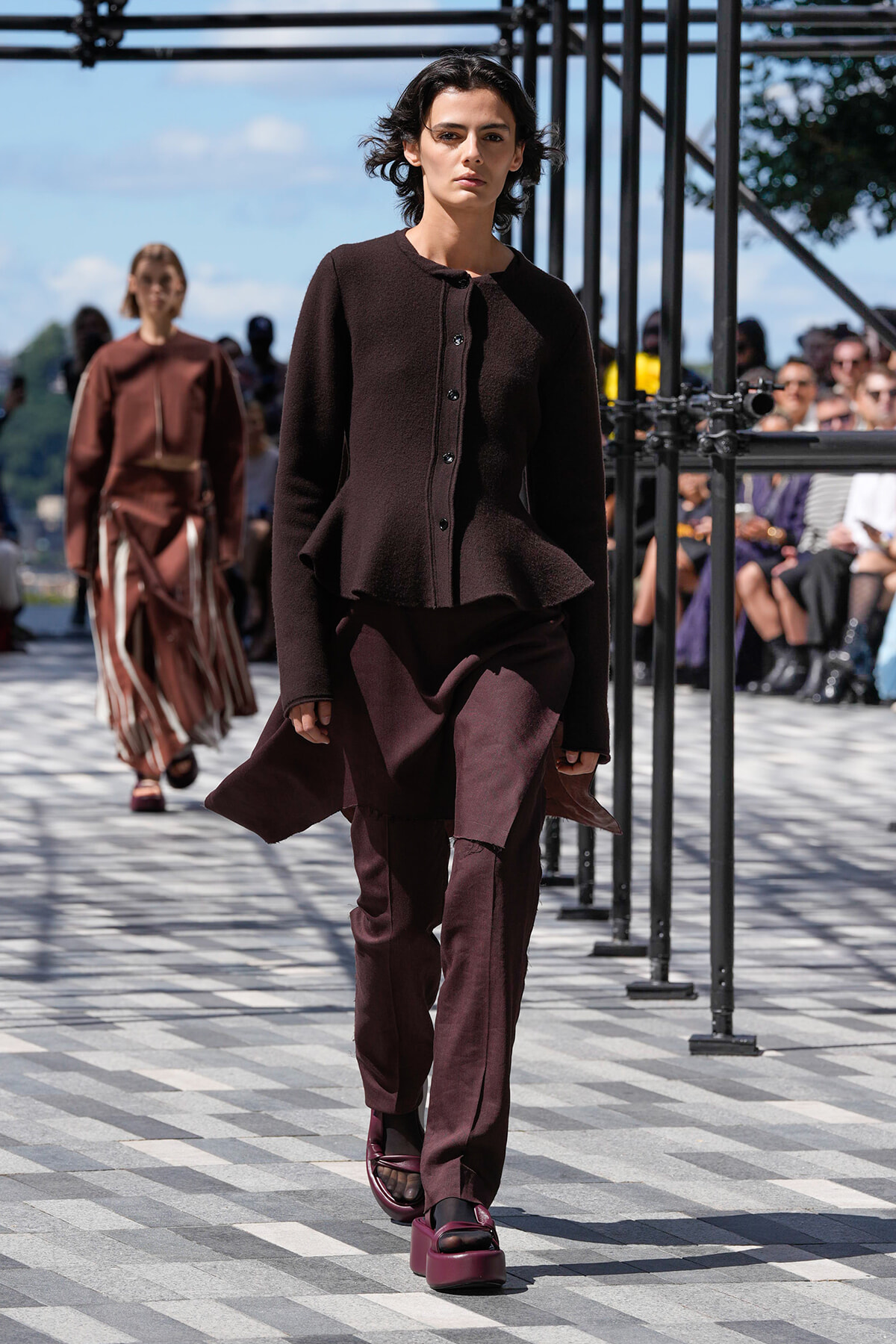 Model walking down a runway in a dark maroon ensemble with a peplum jacket and wide-legged pants; outdoor fashion show with spectators in the background