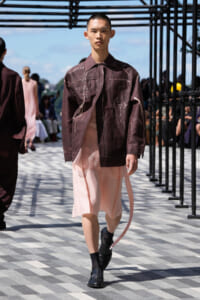 Male fashion model walks the outdoor runway in a dark brown splattered jacket and light pink layered shorts, with black boots.