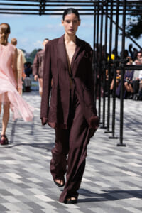 Model walking a dark maroon oversized suit (blazer and wide-leg pants) on a patterned outdoor runway with spectators in the background