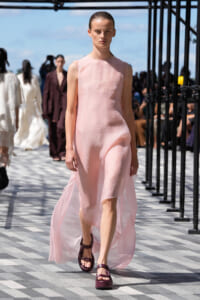 Female model walks a pink sleeveless dress on a daytime runway with burgundy sandals; audience and black metal framework visible in the background.