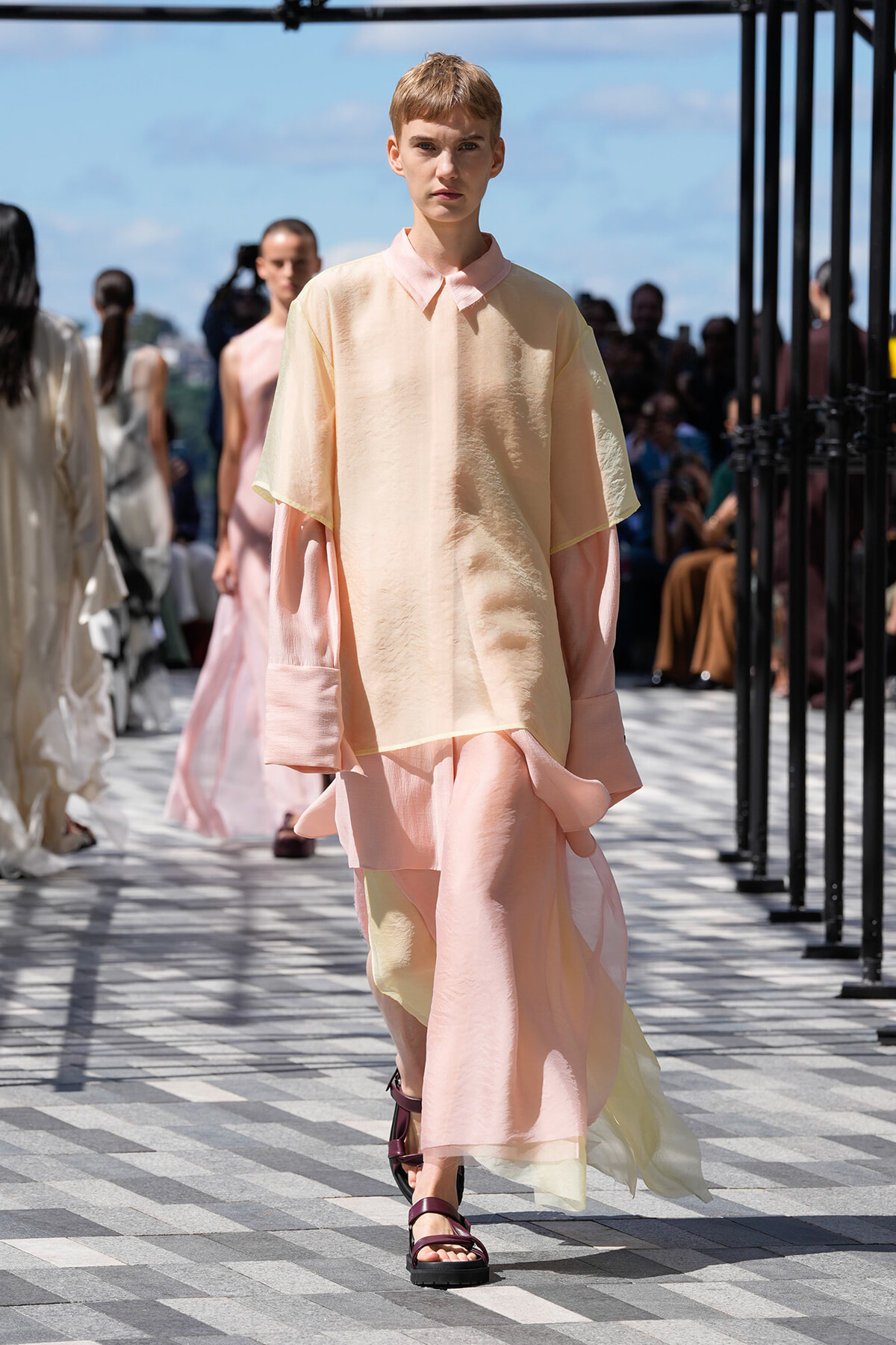 Model walking a runway in pastel peach-pink layered outfit, maroon sandals, during a fashion show with spectators in the background.