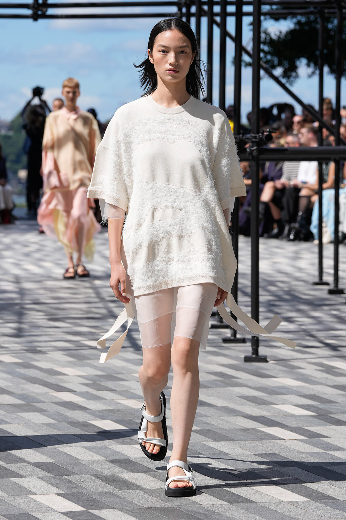 Model walking a runway in a white textured tunic over sheer shorts and white sandals, outdoors on a striped pavement.