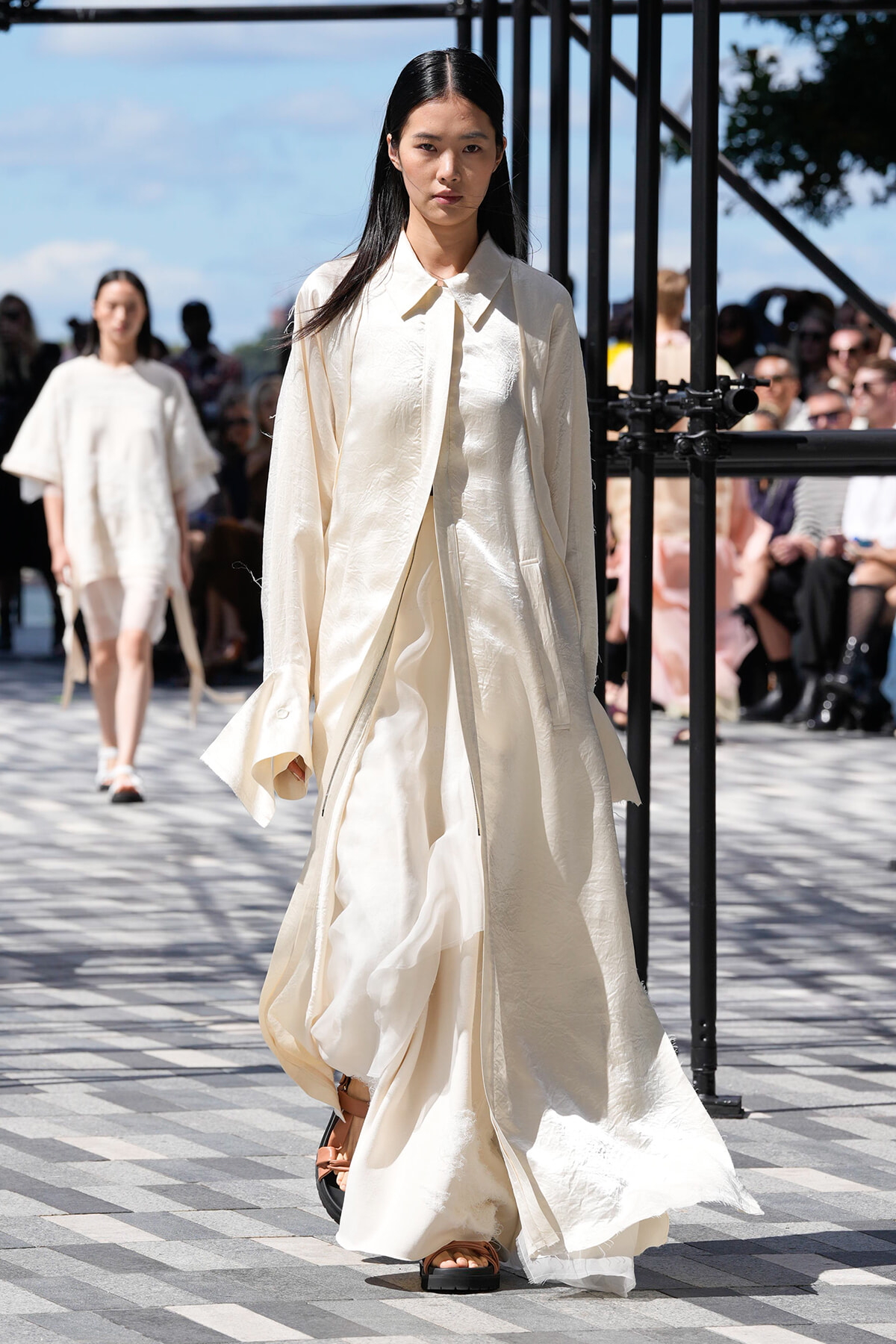 Model walking a runway in a long cream-toned coat with flowing fabric and brown sandals, outdoor fashion show setting.