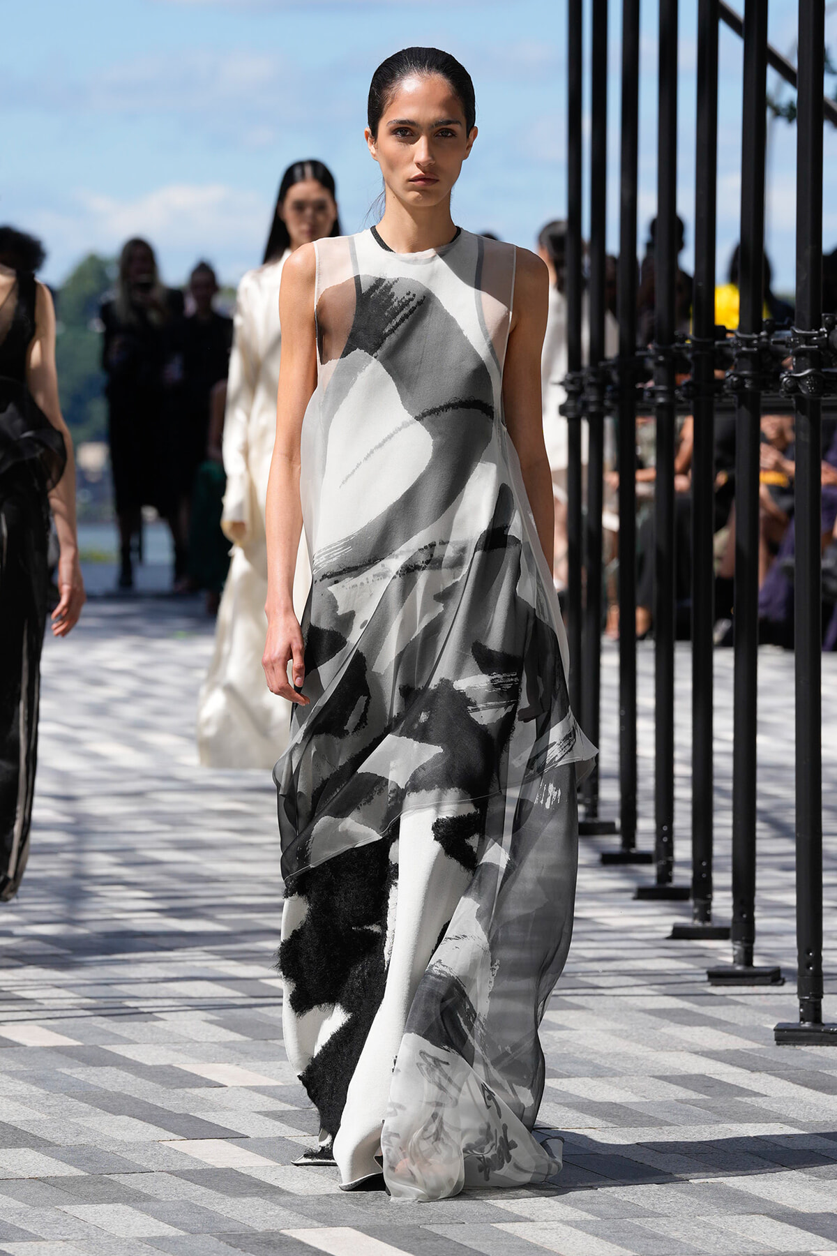 Model walks the runway in a sleeveless, floor-length dress with an abstract black-and-gray brushstroke pattern outdoors.