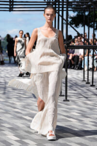 Model in an ivory pleated, layered dress walks a runway outdoors with spectators in the background.