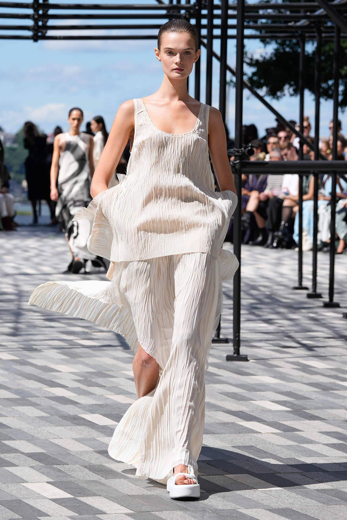 Model in an ivory pleated, layered dress walks a runway outdoors with spectators in the background.