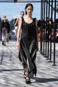 Model walking on an outdoor runway in a black sleeveless top and a black and white striped skirt, wearing platform sandals; blurred audience in the background.