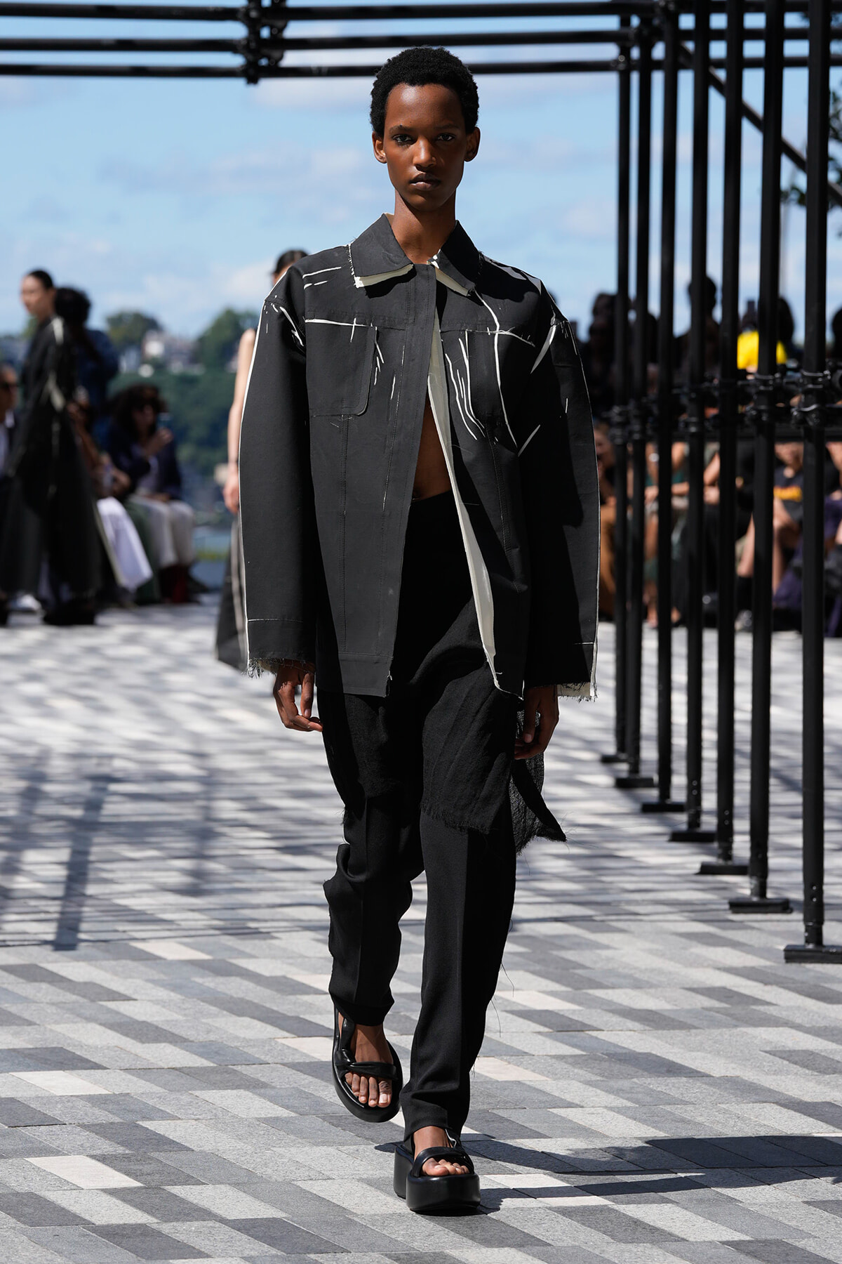 Model walks a dark gray oversized jacket with white streaks on an outdoor runway, paired with black pants and platform sandals.