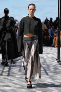 Model walking runway in a cropped black jacket and layered sheer skirt with neutral beige and black panels, outdoor fashion show.