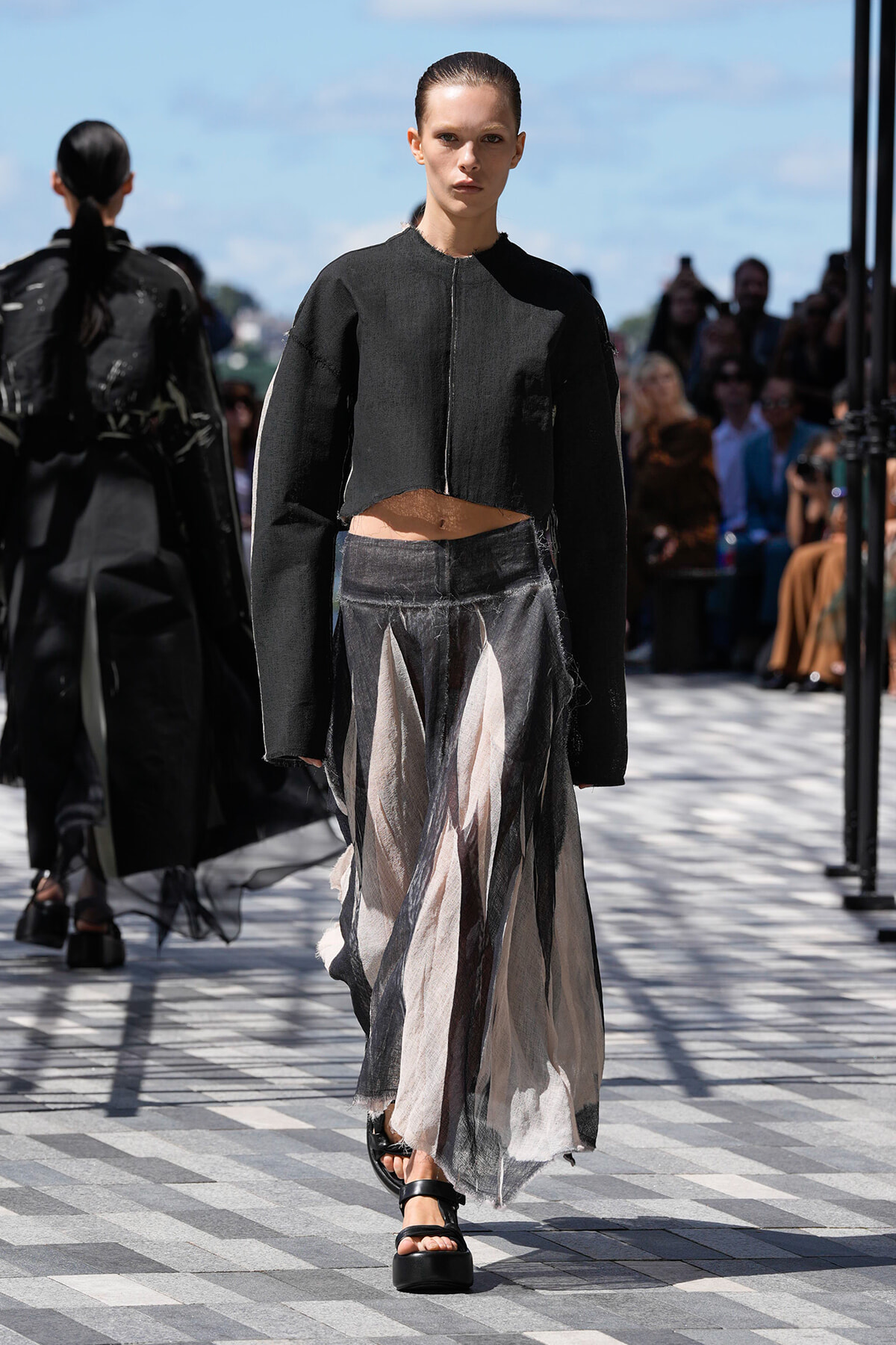 Model walking runway in a cropped black jacket and layered sheer skirt with neutral beige and black panels, outdoor fashion show.