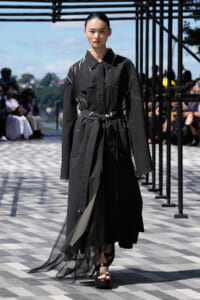 Model walks the runway in a black, paint-splattered long coat with a tied waist belt.