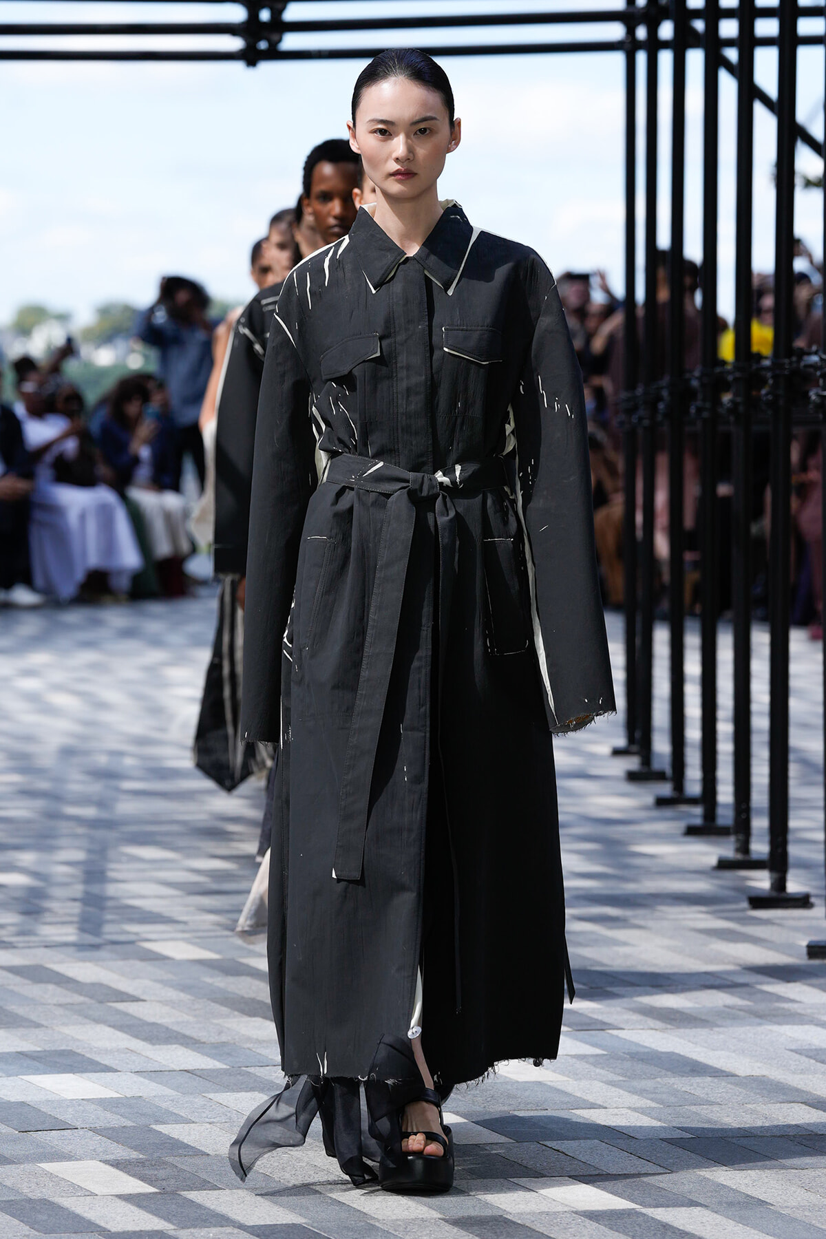 Model walks the runway in a black distressed trench coat with a tied belt and paint-splatter detailing, on a checkered catwalk outdoors.