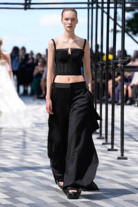 Model walks a black corset-style top and wide-leg fringe pants on an outdoor runway with blurred audience in the background.