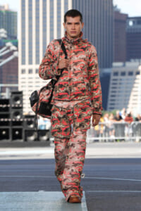 Male model walking on an urban runway in pink camouflage jacket and matching pants, carrying a black shoulder bag and brown boots