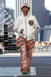 Male model walks runway in a white varsity jacket with patches and pink camouflage pants, urban skyline behind him in a street show setting.