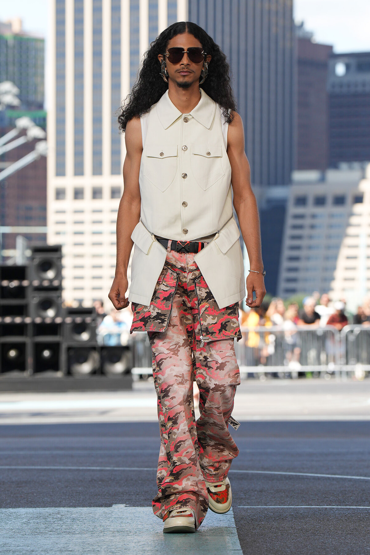 Male model wearing a sleeveless beige utility vest over a shirt, camo cargo pants, and sunglasses on a city runway.