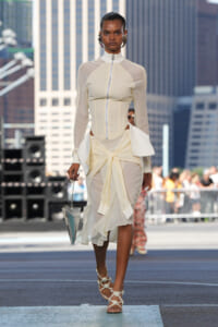 Model in a cream zip-front top with sheer sleeves and a draped ivory skirt on an urban outdoor runway, carrying a geometric bag.