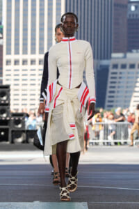 Model walking a street-style runway in a cream outfit with red accents and sandals, city skyline in the background.