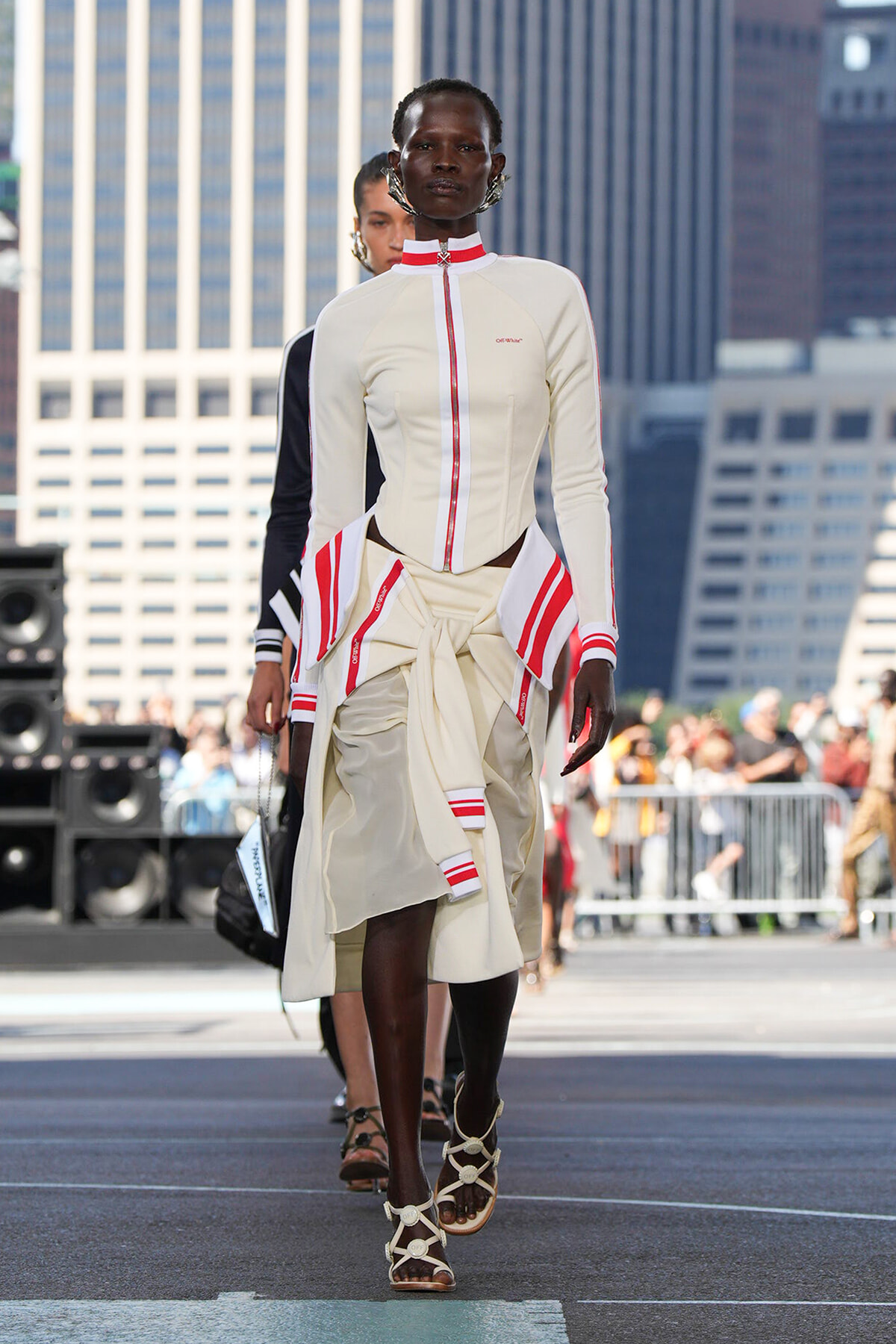 Model walking a street-style runway in a cream outfit with red accents and sandals, city skyline in the background.