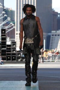 Male model walking a city runway in a black leather sleeveless vest and matching pants with large pocket accents.