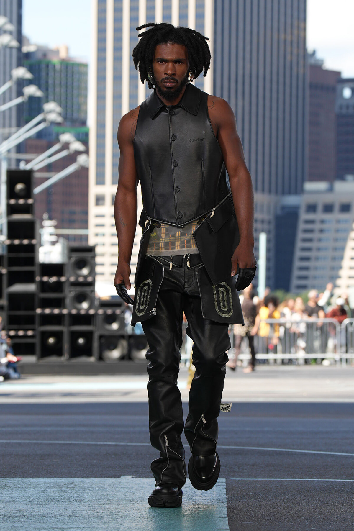 Male model walking a city runway in a black leather sleeveless vest and matching pants with large pocket accents.